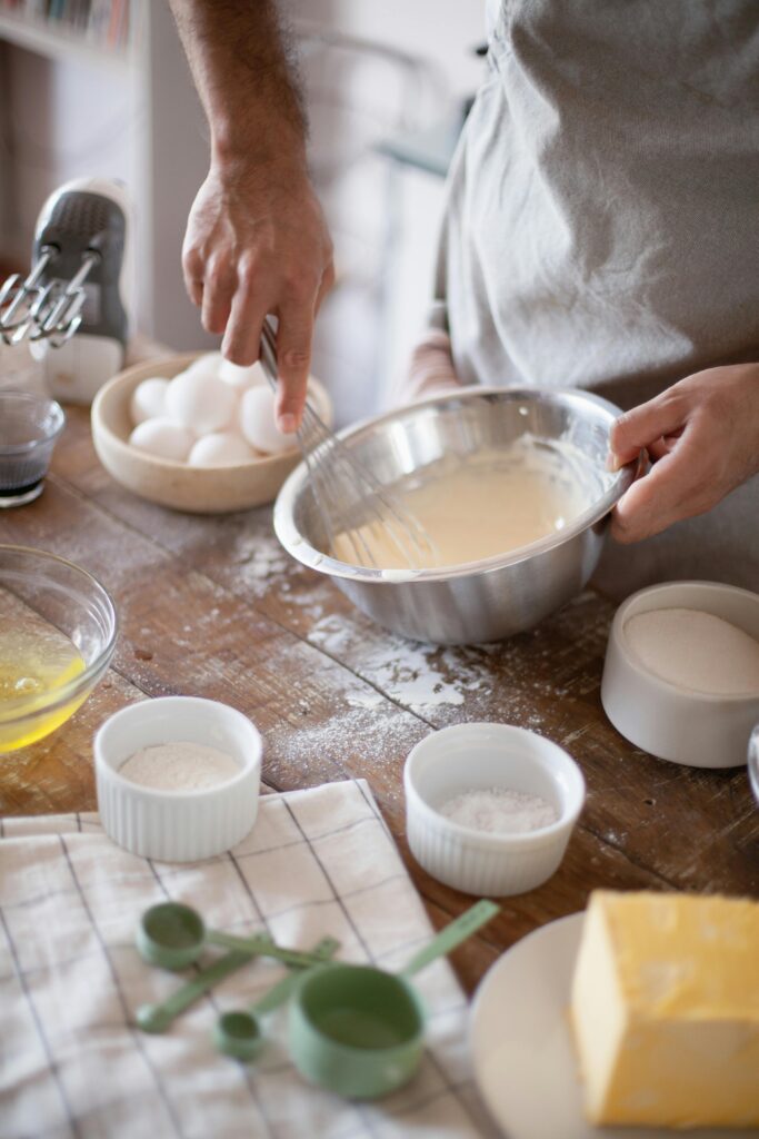 A chef pouring beaten eggs into a mixing bowl while holding a whisk and a bowl of prepared mixture in their hand, preparing ingredients for the recipe.