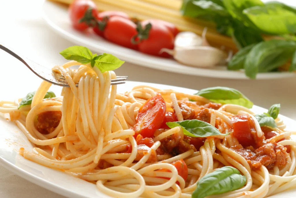 A close-up of spaghetti carbonara dish topped with a rich tomato sauce, cherry tomatoes, fresh basil leaves, and a fork twirling the pasta. A plate of fresh ingredients, including tomatoes, garlic, basil.