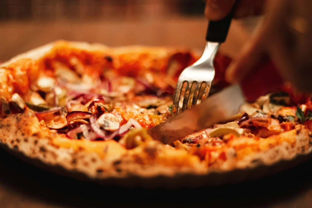 "Close-up of a freshly baked pizza with a golden crust, topped with red onions, mushrooms, and olives, as a fork and knife cut through a slice."