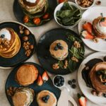 Delicious stacks of pancakes topped with fresh blueberries, strawberries, walnuts, and yogurt, surrounded by spinach leaves and chocolate drizzle, perfect for a sweet morning treat.