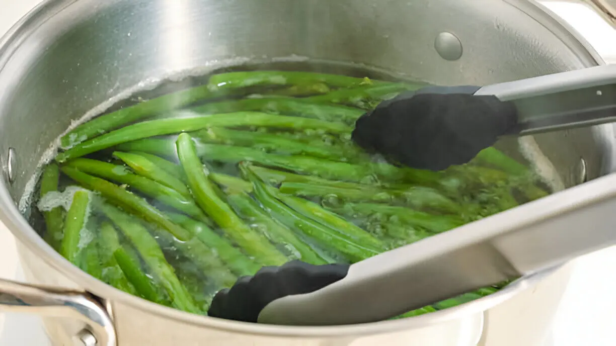 Fresh green beans being blanched in boiling water inside a stainless steel pot, held with black tongs.