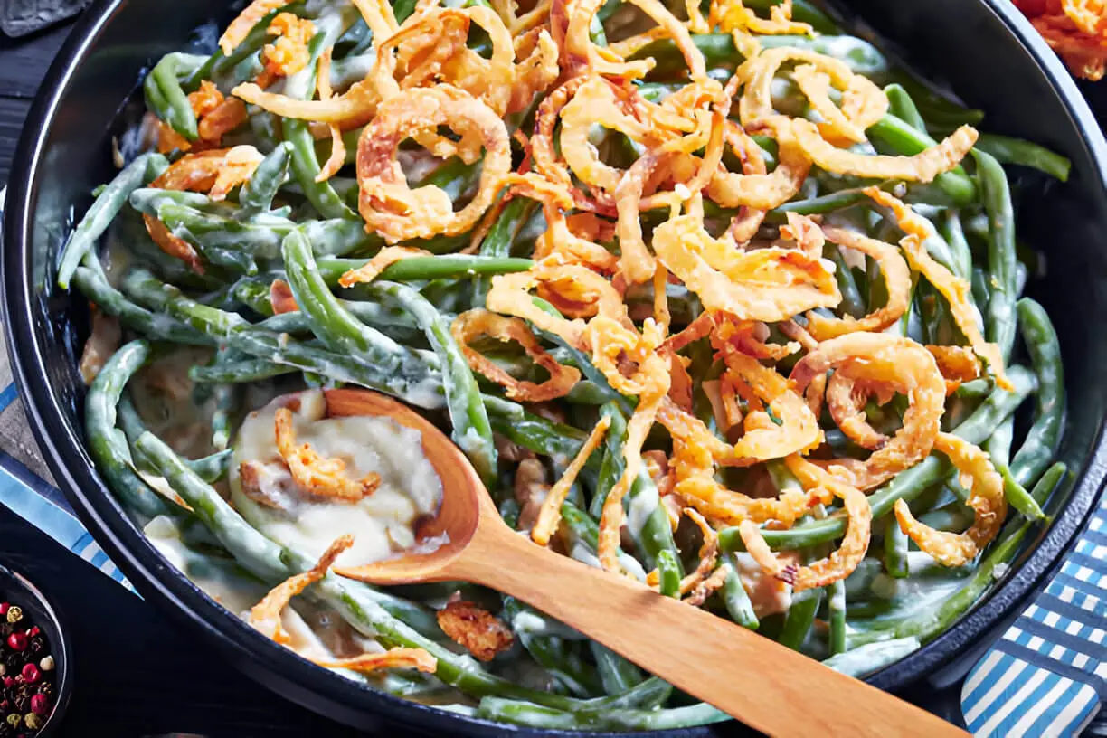 Fresh Green Bean Casserole in a black dish topped with crispy onion rings, served with a wooden spoon, ideal for festive gatherings.