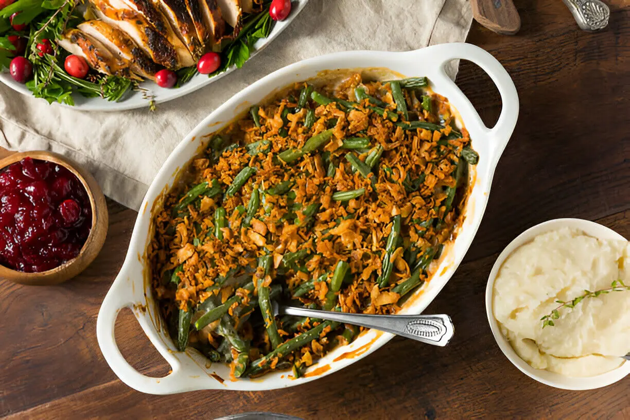 A casserole dish filled with green bean casserole topped with crispy onions, surrounded by side dishes like mashed potatoes, cranberry sauce, and roasted turkey slices on a wooden table.