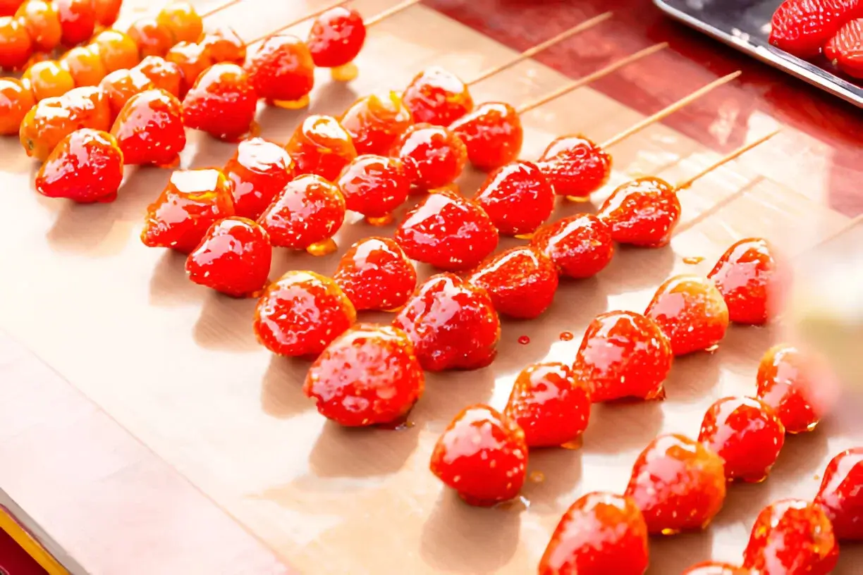 Glossy Tanghulu skewers made with strawberries and golden fruits, displayed on a parchment-lined tray under warm light.